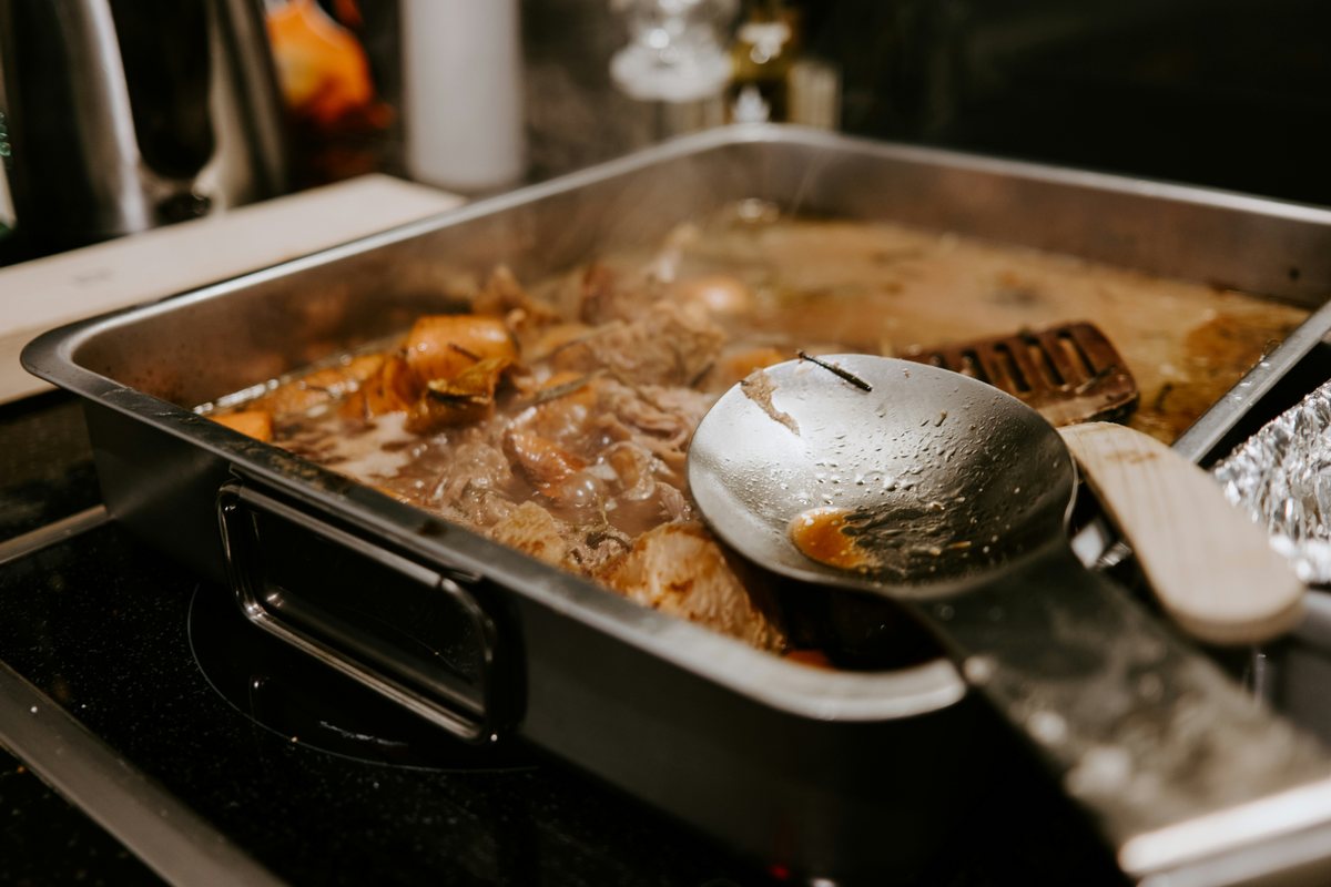 Cooked food and its juices in a metal roasting tin beside a serving spoon.