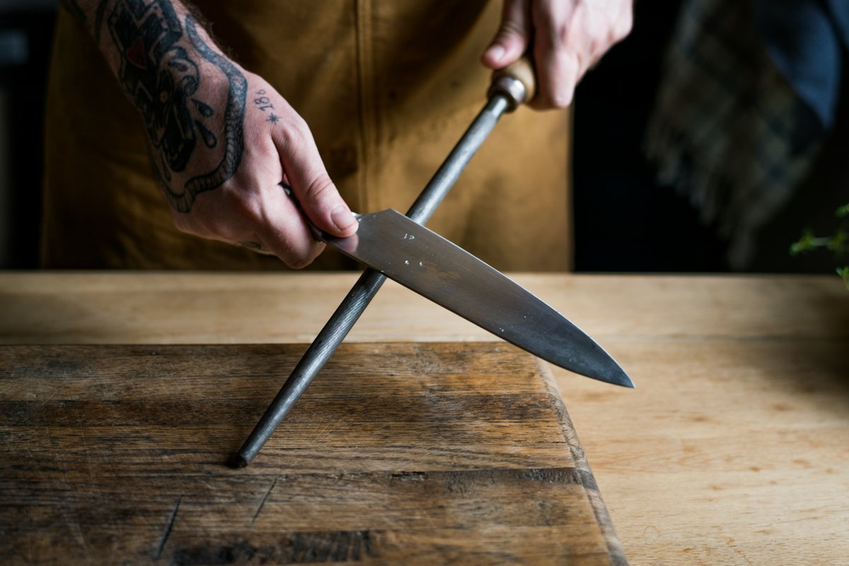 A cook holding a kitchen knife against a honing rod with its tip resting on a board.