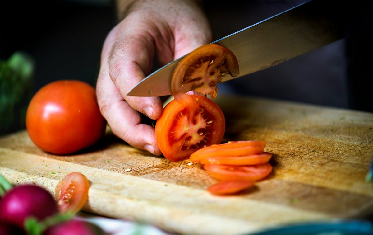 A hand using a kitchen knife to slice a ripe tomato on a wooden board.