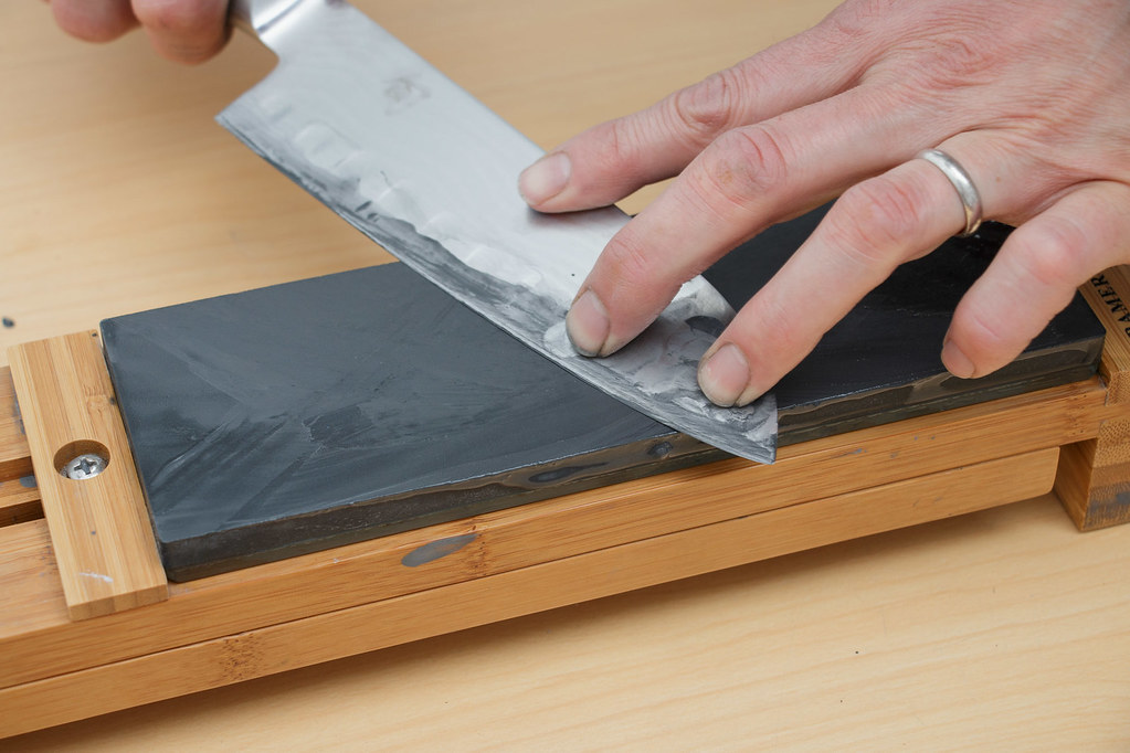 Hands holding a kitchen knife at an angle against a whetstone on a wooden base.
