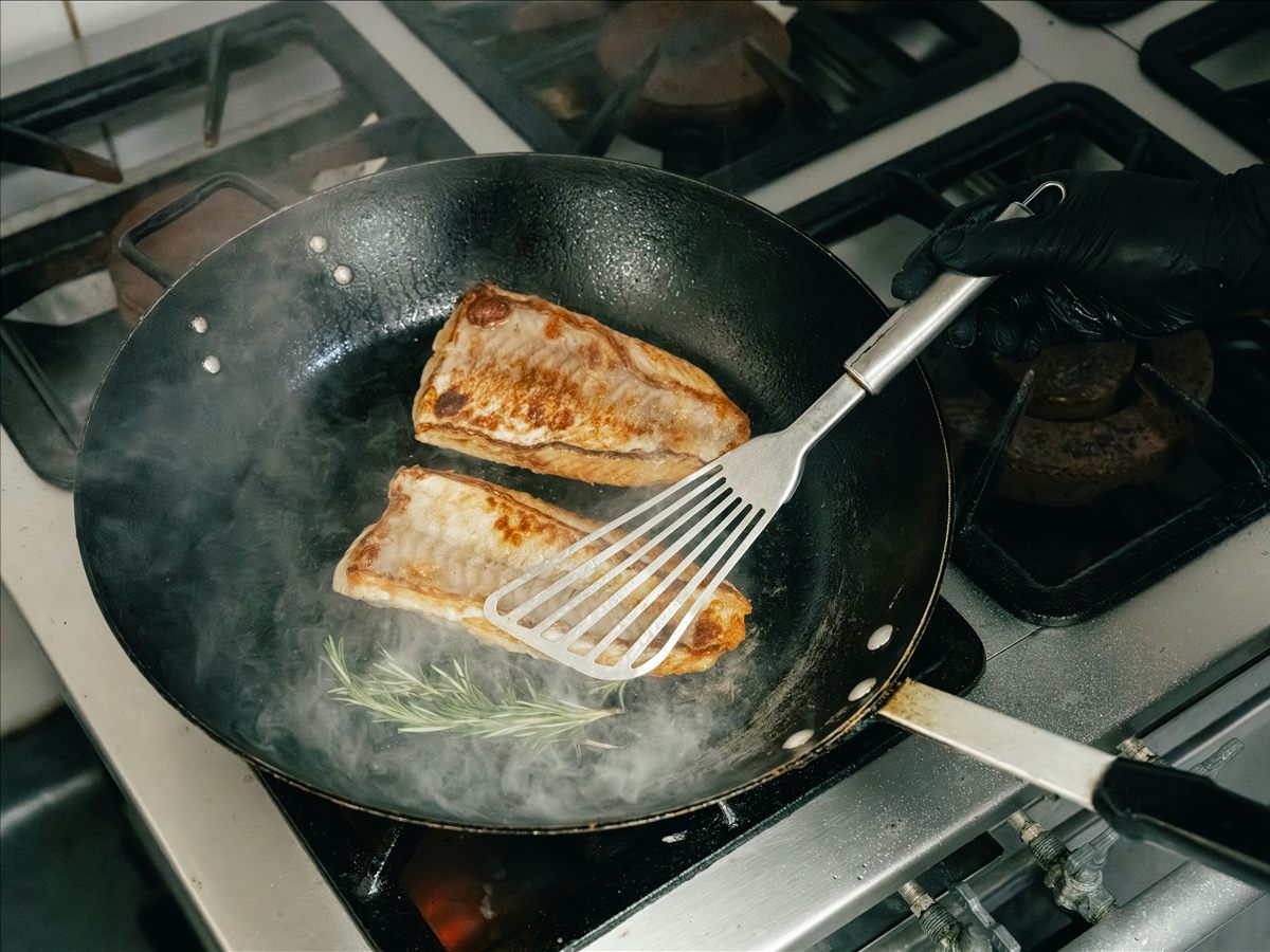 A cook lifting browned fish fillets from a steaming pan with a spatula.