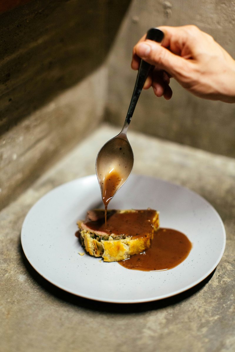 A hand spooning brown sauce over a plated savoury dish.