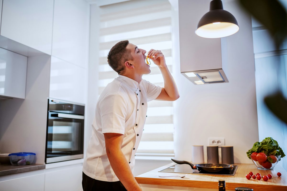 A chef tasting a small bite while cooking beside a pan in a bright kitchen.