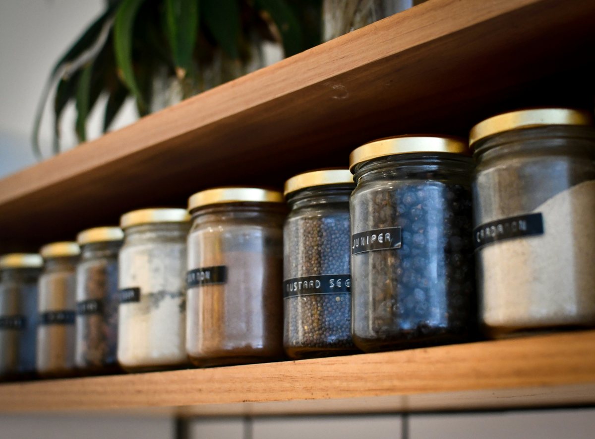 Glass jars of spices, including mustard seed and juniper, lined up on a wooden shelf.