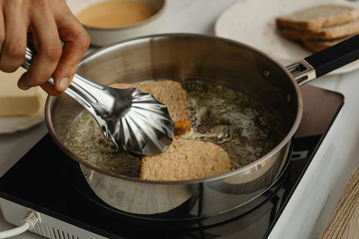 A hand using tongs to turn pieces of bread in a stainless steel frying pan.
