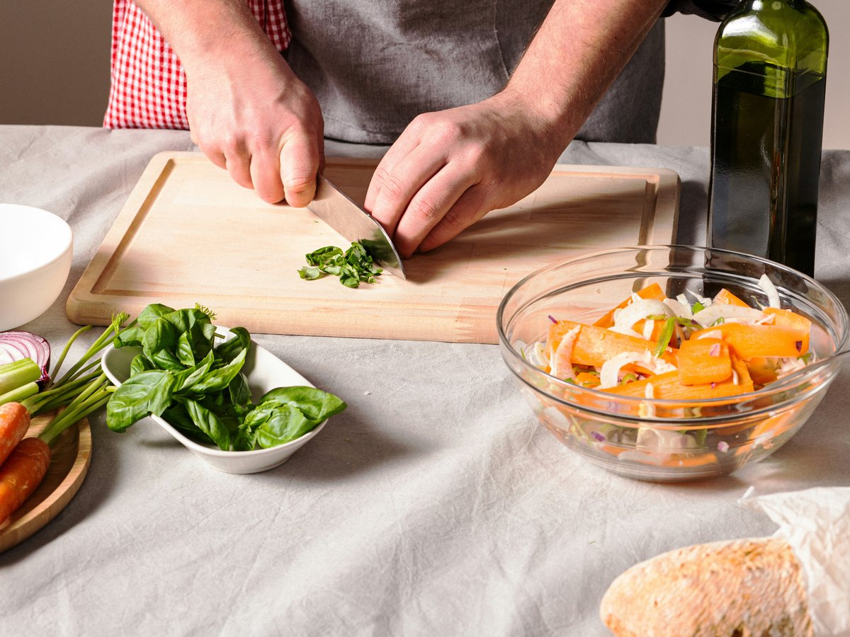 A cook cuts basil leaves with a knife on a wooden board.