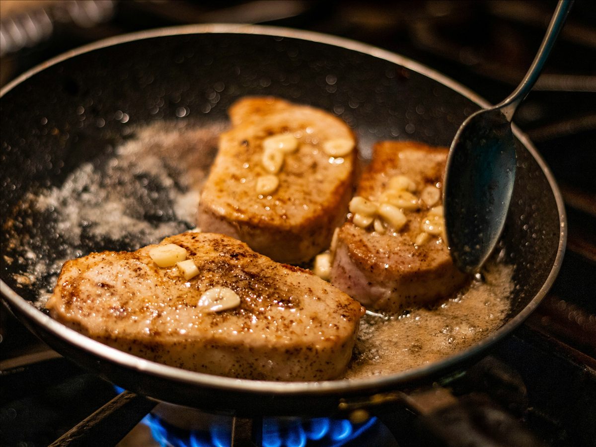 Three pork chops browning with garlic in a dark pan over a gas flame.