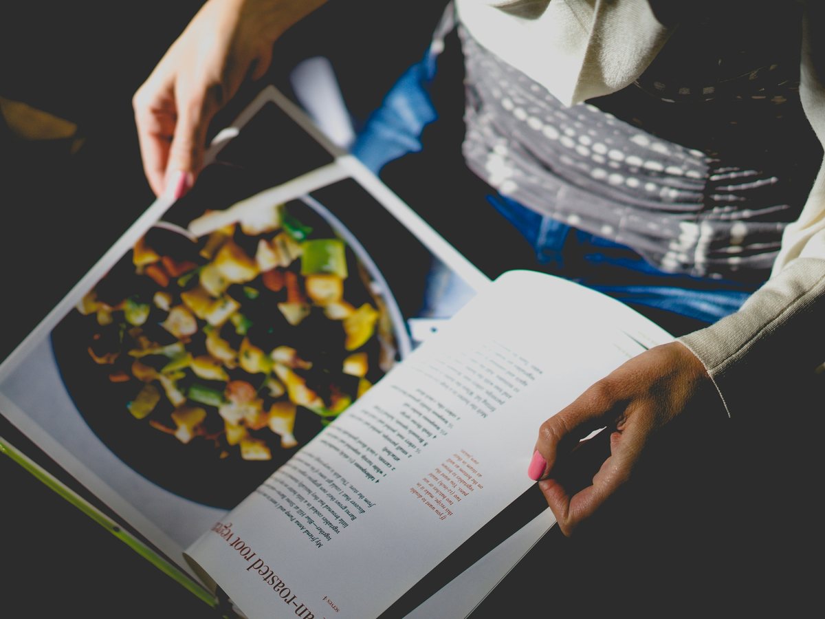 A person holding an open cookbook with a food photograph and printed recipe.